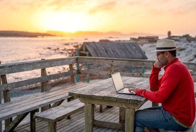 A man on a beach at sunset looking at his laptop instead of the sunset.