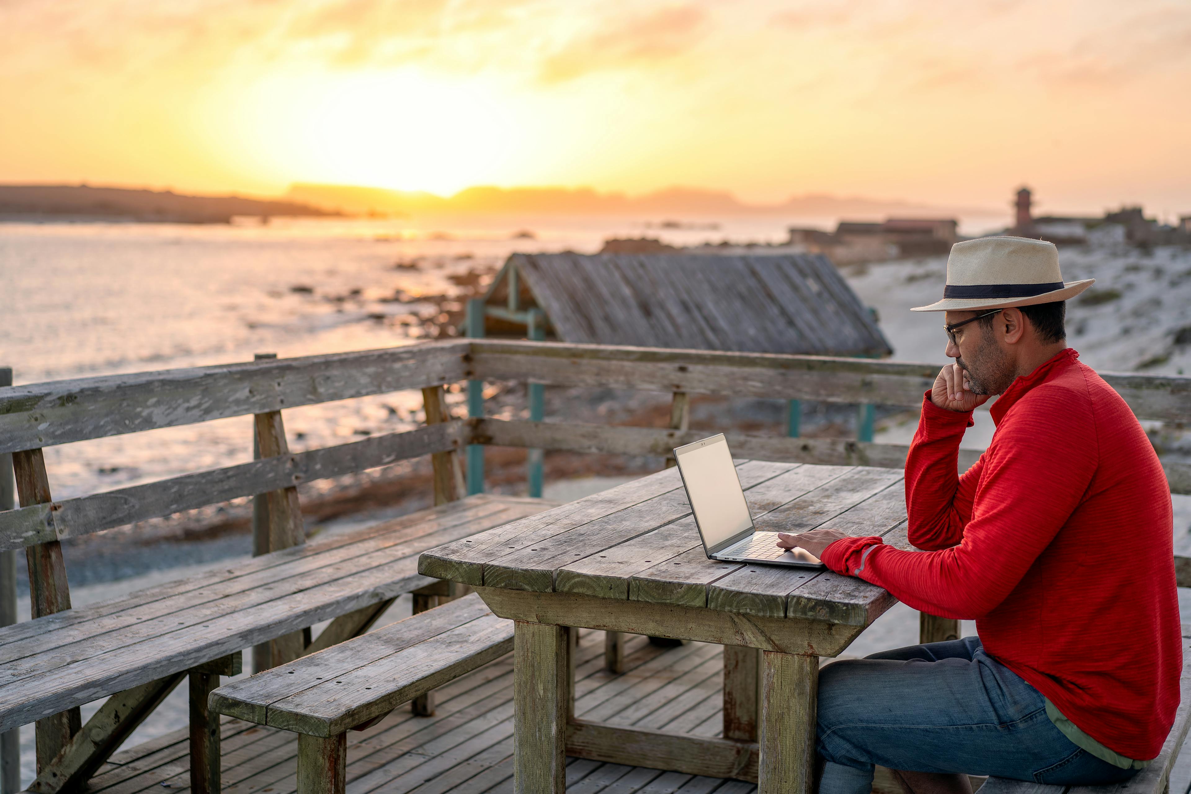 A man on a beach at sunset looking at his laptop instead of the sunset.